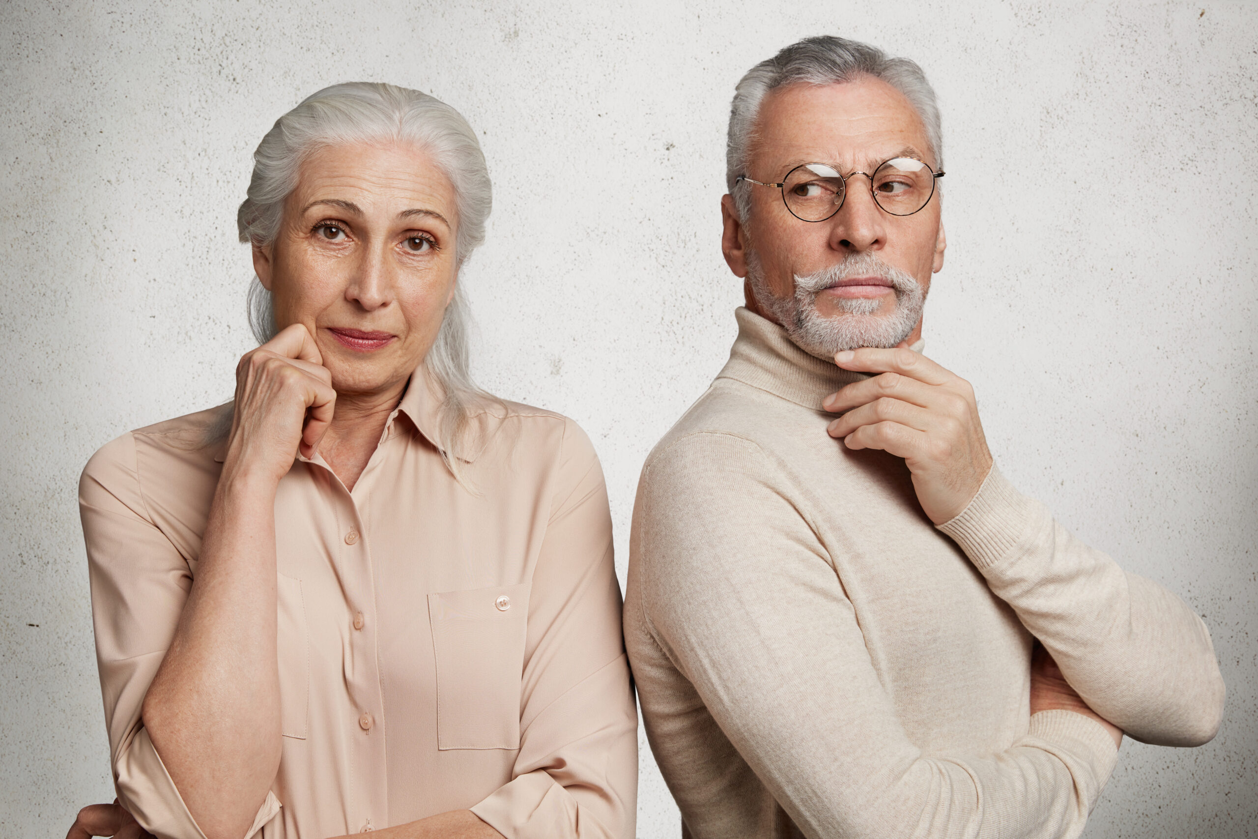 Mature couple poses against white concrete wall. Pretty elederly woman in beige blouse and makeup, her husband stands back, looks thoughtfully aside, being on pension, spend free time together